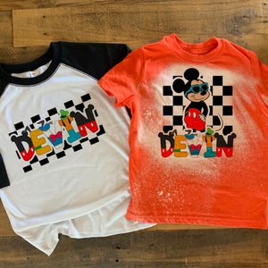 May include: Two t-shirts with a bleach-splattered design featuring Mickey Mouse and the name "DEVIN" in colorful letters. The shirts are on a wooden surface.