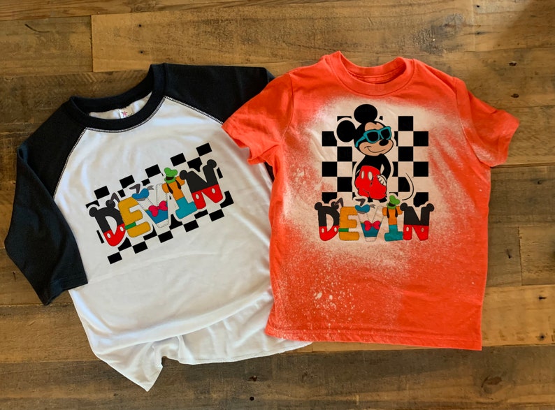 May include: Two t-shirts with a bleach-splattered design featuring Mickey Mouse and the name "DEVIN" in colorful letters. The shirts are on a wooden surface.
