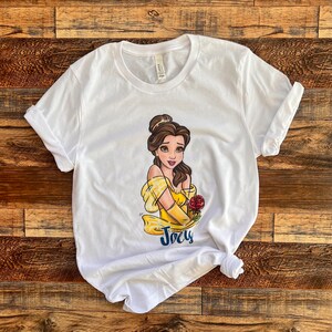 Peut inclure: T-shirt blanc avec une image de dessin anim&eacute; de Belle de La Belle et la B&ecirc;te. Belle porte une robe jaune et tient une rose rouge. Le t-shirt porte le nom "Joely" imprim&eacute; sous l'image.