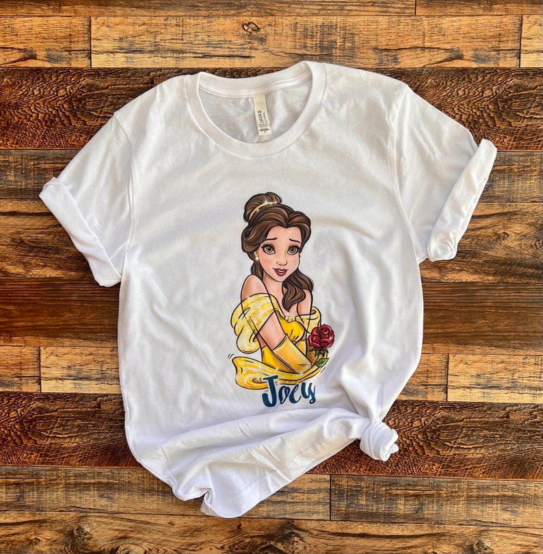 Peut inclure: T-shirt blanc avec une image de dessin anim&eacute; de Belle de La Belle et la B&ecirc;te. Belle porte une robe jaune et tient une rose rouge. Le t-shirt porte le nom "Joely" imprim&eacute; sous l'image.