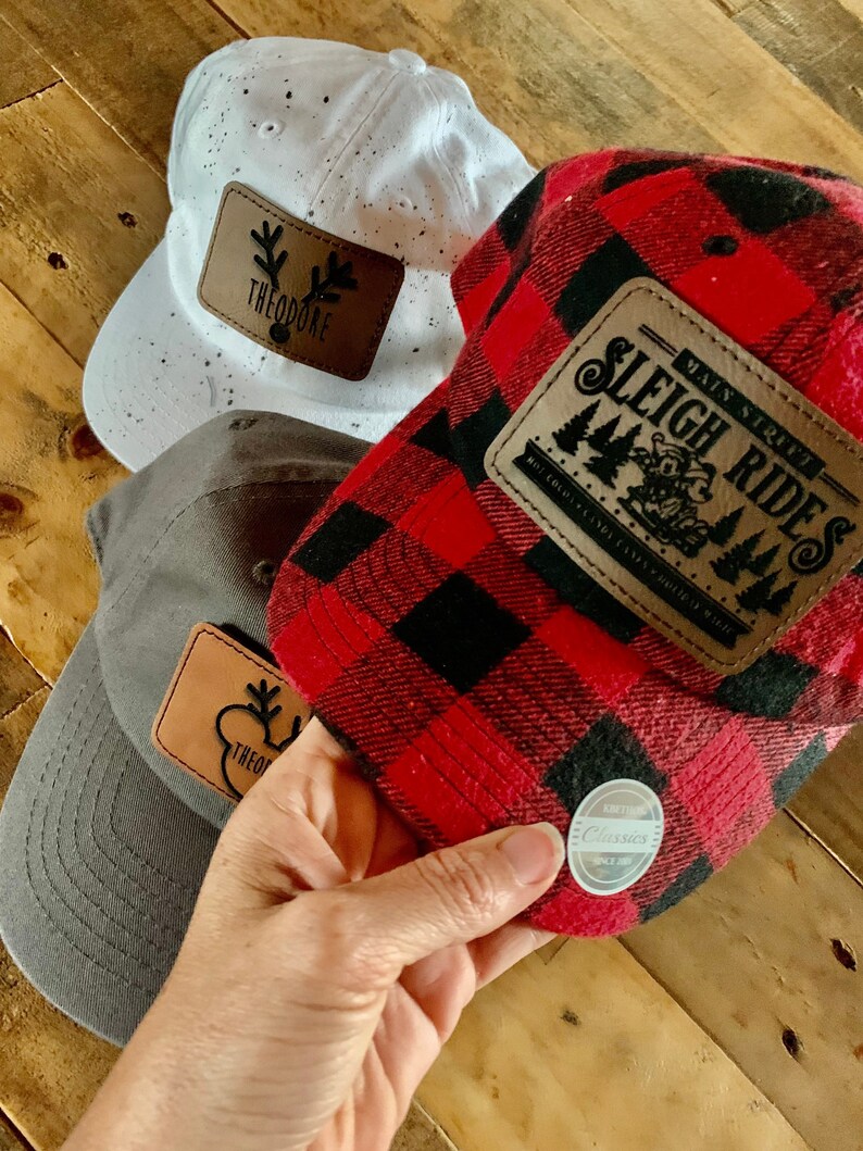 May include: Three baseball caps with different designs. One is white with black speckles and a brown leather patch that says "Theodore". Another is red and black plaid with a brown leather patch that says "Main Street Sleigh Rides". The third is gray with a brown leather patch that says "Theodore".