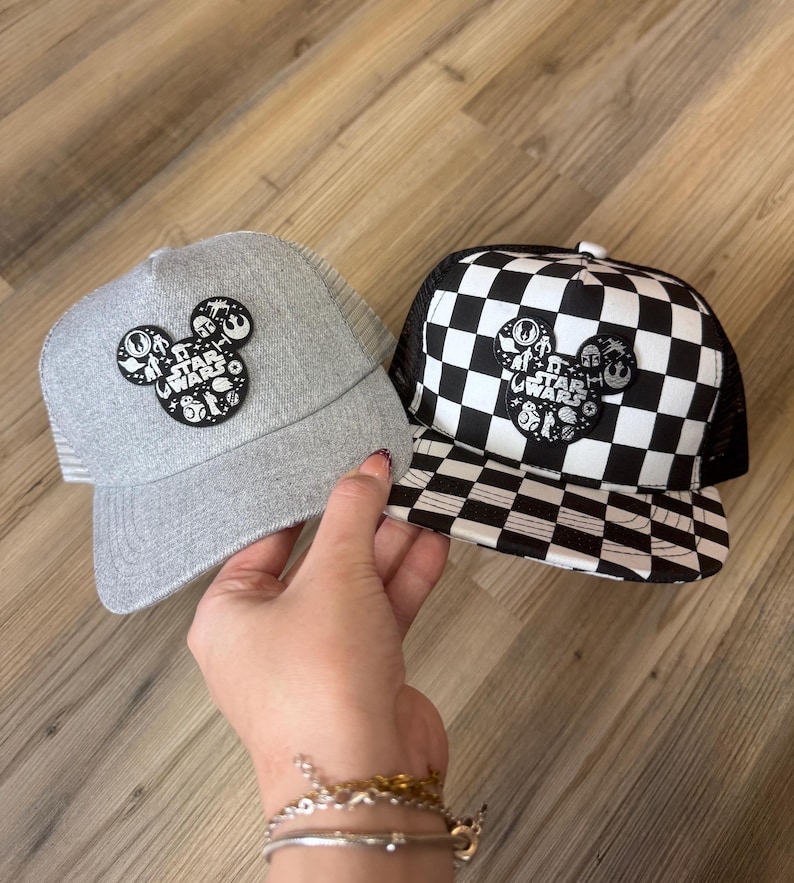 May include: Two baseball caps with a Star Wars theme. One is grey with a mesh back and a Mickey Mouse-shaped patch. The other is black and white chequered with a mesh back and a similar patch. Both patches feature Star Wars characters.