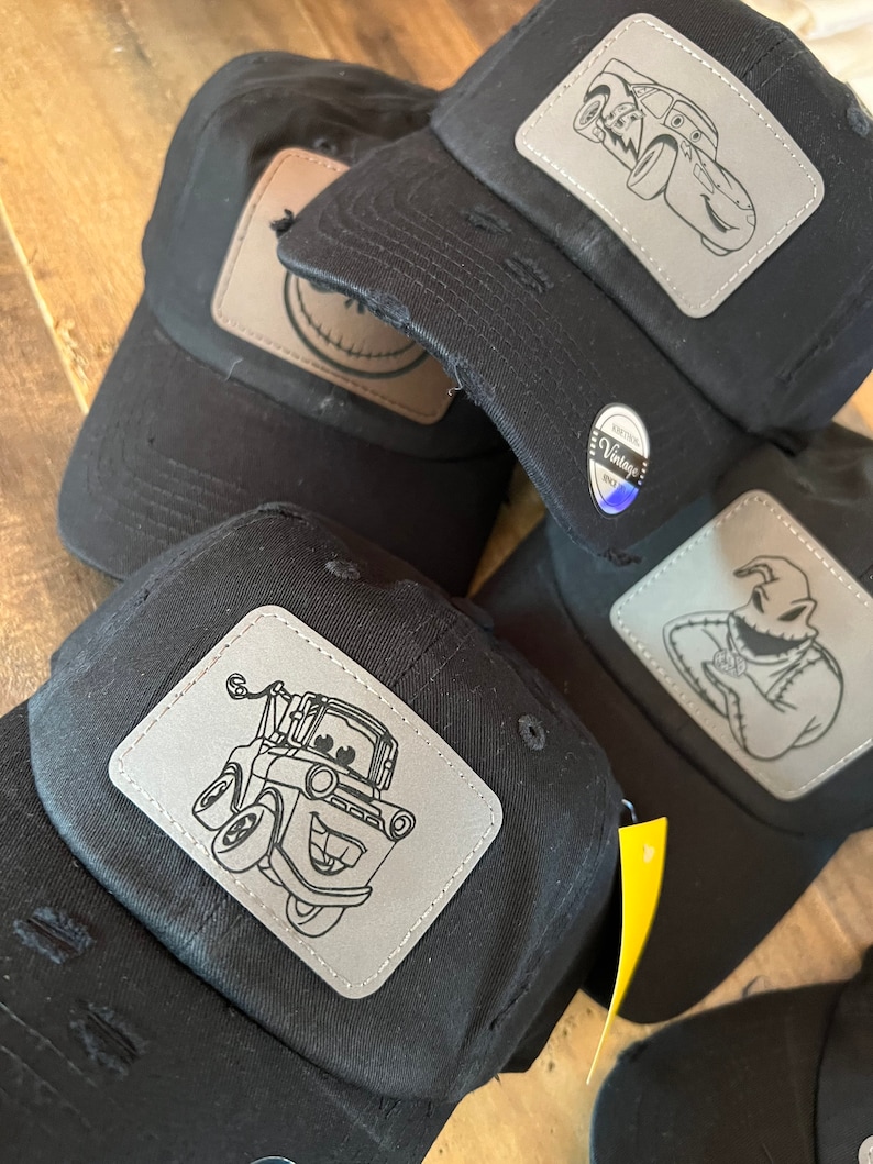 May include: Four black baseball caps with distressed fabric and different character patches. The patches are gray and feature a tow truck, a race car, a ghost, and a smiley face.
