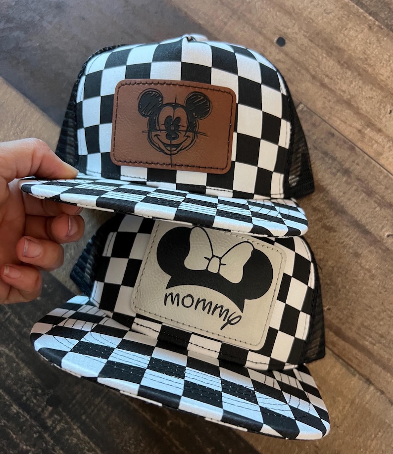 K&ouml;nnte beinhalten: Zwei Trucker-H&uuml;te mit einem schwarz-wei&szlig;en Schachbrettmuster. Einer der H&uuml;te hat einen braunen Lederaufn&auml;her mit einer Mickey-Mouse-Skizze, der andere einen wei&szlig;en Lederaufn&auml;her mit einer Minnie-Mouse-Silhouette und dem Wort "mommp".