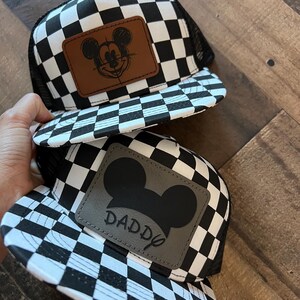 K&ouml;nnte beinhalten: Zwei schwarz-wei&szlig; karierte Trucker-H&uuml;te. Ein Hut hat einen braunen Lederaufn&auml;her mit einer Mickey-Mouse-Skizze. Der andere Hut hat einen grauen Lederaufn&auml;her mit einer Mickey-Mouse-Silhouette und dem Wort "DADDY".