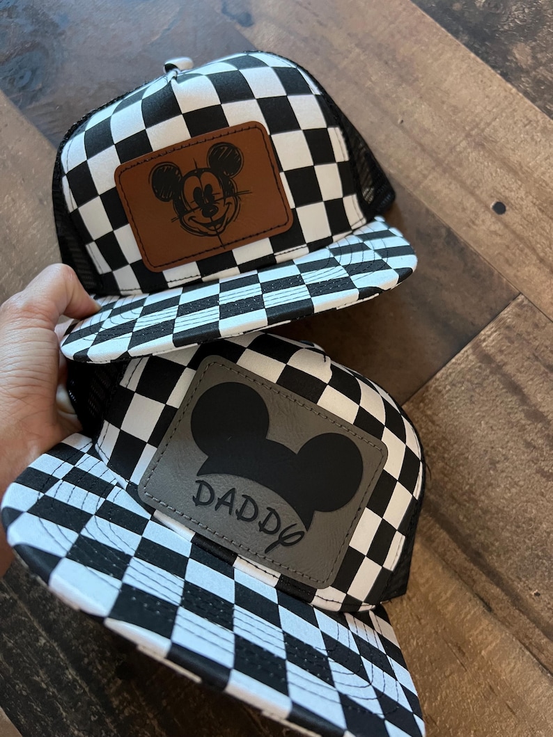K&ouml;nnte beinhalten: Zwei schwarz-wei&szlig; karierte Trucker-H&uuml;te. Ein Hut hat einen braunen Lederaufn&auml;her mit einer Mickey-Mouse-Skizze. Der andere Hut hat einen grauen Lederaufn&auml;her mit einer Mickey-Mouse-Silhouette und dem Wort "DADDY".