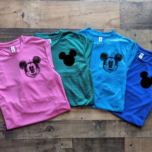 May include: Four colorful t-shirts: pink, teal, blue, and royal blue. Each shirt features a Mickey Mouse graphic or silhouette. The Mickey Mouse graphic is a black sketch, while the silhouette is solid black.