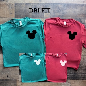 May include: Four short-sleeved shirts in teal and red, each with a Mickey Mouse head silhouette on the chest. The shirts are displayed on a wooden surface. The text "DRI FIT" is at the top.
