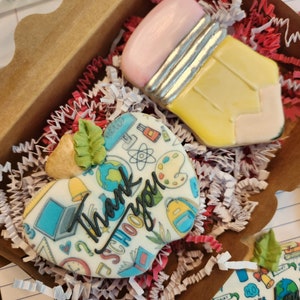 May include: A gift box containing decorated sugar cookies. One cookie is shaped like a yellow pencil with a pink eraser. Another cookie is heart-shaped with the words "Thank you SCHOOL" and school-themed illustrations.