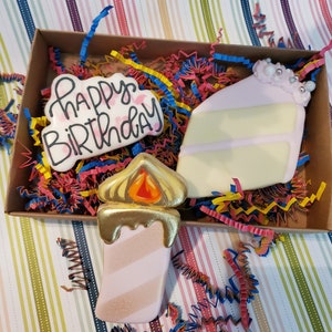May include: A gift box with three decorated sugar cookies. One is a candle with gold detailing and a flame. Another is a cake slice with pink icing. The third cookie says "Happy Birthday". The box is filled with colourful paper shreds.
