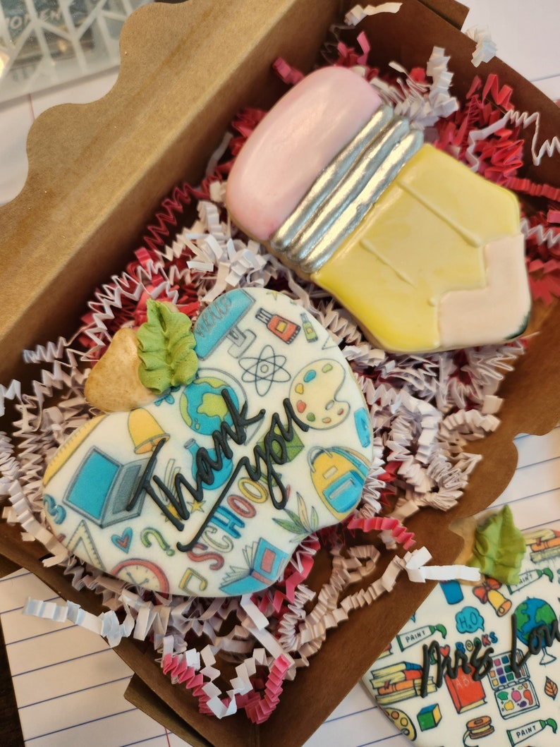 May include: A gift box containing decorated sugar cookies. One cookie is shaped like a yellow pencil with a pink eraser. Another cookie is heart-shaped with the words "Thank you SCHOOL" and school-themed illustrations.