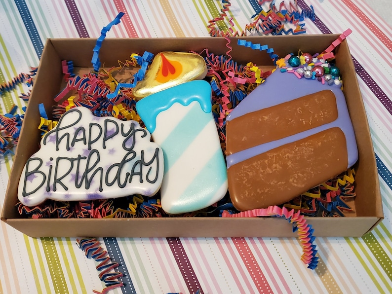 May include: A gift box with decorated sugar cookies. The cookies feature a candle, a cake slice, and a cookie with the words "Happy Birthday". The cookies are decorated with blue, white, purple, and brown icing. The box is filled with colourful paper shreds.