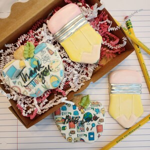 May include: A wooden box filled with red and white paper shreds, holding decorated sugar cookies. The cookies are shaped like pencils and apples, with the words "Thank you SCHOOL" and "Miss Love" on the apple cookies. Yellow pencils are scattered around.
