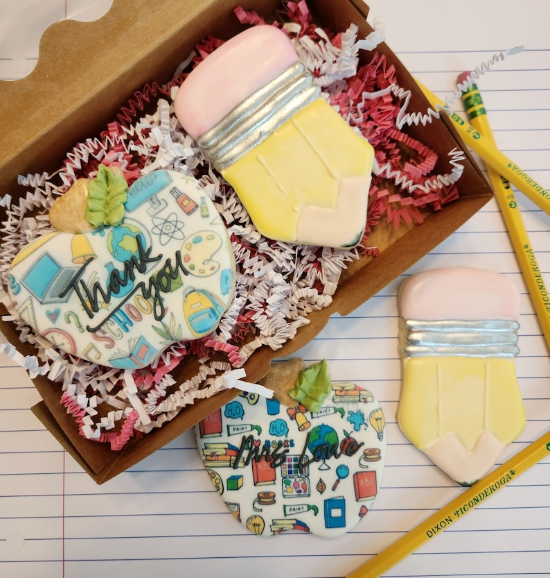 May include: A wooden box filled with red and white paper shreds, holding decorated sugar cookies. The cookies are shaped like pencils and apples, with the words "Thank you SCHOOL" and "Miss Love" on the apple cookies. Yellow pencils are scattered around.