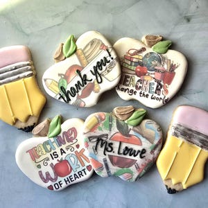 May include: Assortment of decorated sugar cookies in the shape of pencils and apples. The apple cookies have various teacher-themed messages, including "thank you!" and "Teachers change the world."