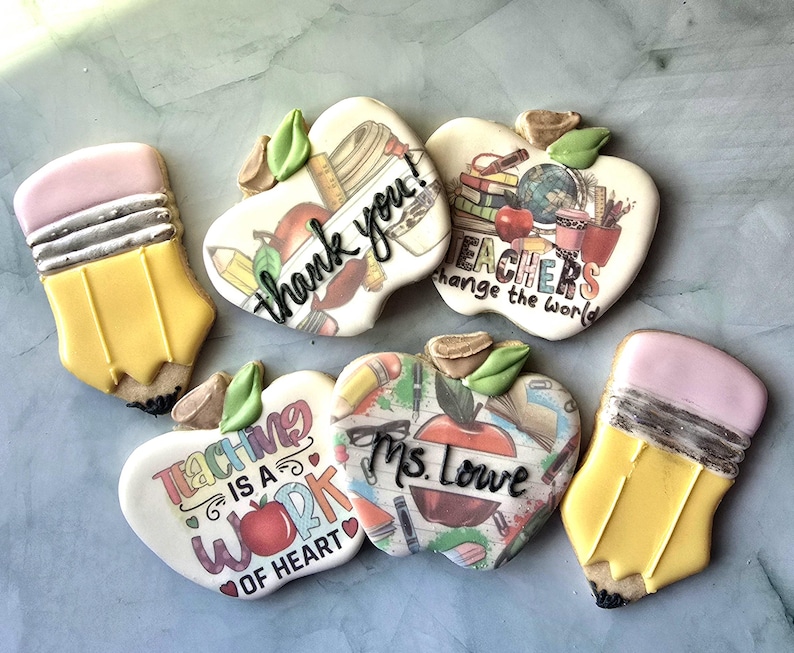 May include: Assortment of decorated sugar cookies in the shape of pencils and apples. The apple cookies have various teacher-themed messages, including "thank you!" and "Teachers change the world."