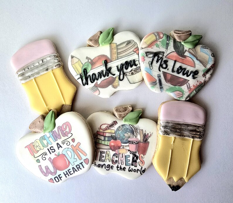 May include: Assortment of decorated sugar cookies in the shape of pencils and apples. The apple cookies have messages like "thank you", "Ms. Lowe", and "Teaching is a work of heart". The cookies are decorated with edible ink.