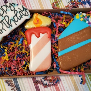 May include: A box of decorated sugar cookies with a birthday theme. The cookies include a cloud-shaped cookie with "Happy Birthday" written on it, a candle, and a slice of cake with blue icing. The box is filled with colourful paper shreds.