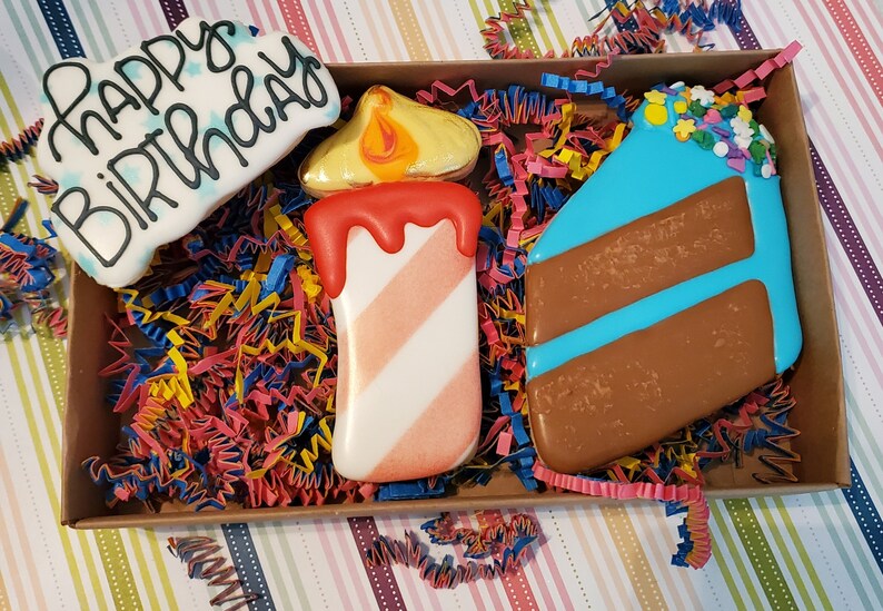 May include: A box of decorated sugar cookies with a birthday theme. The cookies include a cloud-shaped cookie with "Happy Birthday" written on it, a candle, and a slice of cake with blue icing. The box is filled with colourful paper shreds.