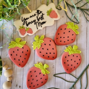 May include: Six decorated sugar cookies shaped like strawberries. The cookies are red with black seeds and green leaves. One cookie has the text "You are the berry best Mom" in black lettering.