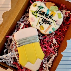 May include: A gift box filled with two decorated sugar cookies. One cookie is shaped like a yellow pencil with a pink eraser and silver band. The other is a heart-shaped cookie with the words "Thank you" and school-themed illustrations.