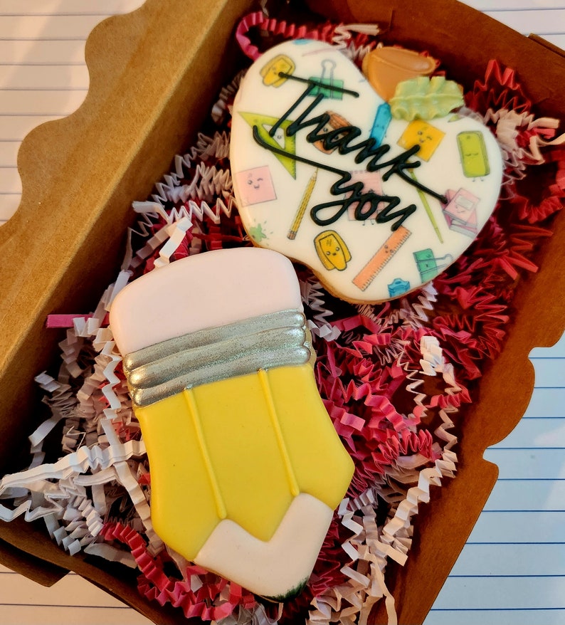 May include: A gift box filled with two decorated sugar cookies. One cookie is shaped like a yellow pencil with a pink eraser and silver band. The other is a heart-shaped cookie with the words "Thank you" and school-themed illustrations.
