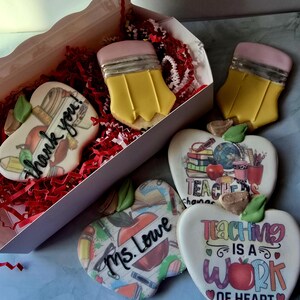 May include: A collection of decorated sugar cookies in the shape of pencils and apples. The cookies are yellow, pink, white, and red. Some cookies have the words "Thank you!", "Ms. Lowe", and "Teaching is a work of heart" on them. The cookies are in a white box with red paper.