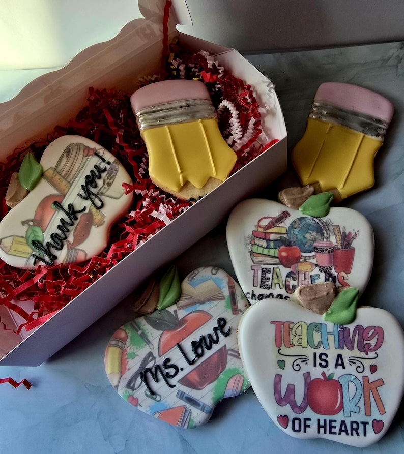 May include: A collection of decorated sugar cookies in the shape of pencils and apples. The cookies are yellow, pink, white, and red. Some cookies have the words "Thank you!", "Ms. Lowe", and "Teaching is a work of heart" on them. The cookies are in a white box with red paper.