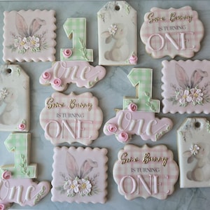 May include: A collection of decorated sugar cookies with a bunny theme. The cookies are in various shapes, including squares and tags, and feature bunny ears, floral designs, and the text "Some Bunny is Turning One."