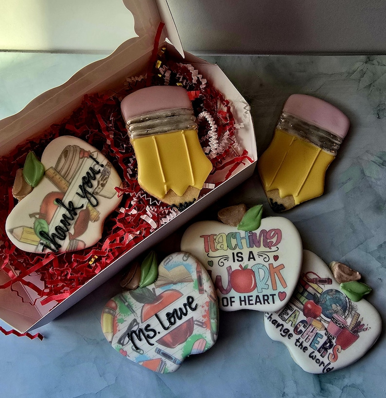 May include: A collection of decorated sugar cookies in a gift box. The cookies are shaped like pencils and apples. Some cookies have text that says "thank you", "Teaching is a work of heart", "Ms. Lowe", and "Teachers change the world".