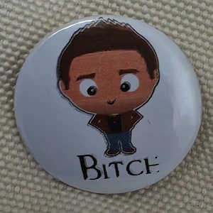 May include: A white button badge with a cartoon illustration of a person with brown hair, a brown jacket, and blue jeans. The word "Bitch" is printed in black at the bottom of the badge.