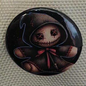 May include: A circular, glossy button with a spooky doll design. The doll has stitched features, including cross-shaped eyes and a stitched mouth. It wears a hooded cloak and a red bow. The background is black with blue accents.