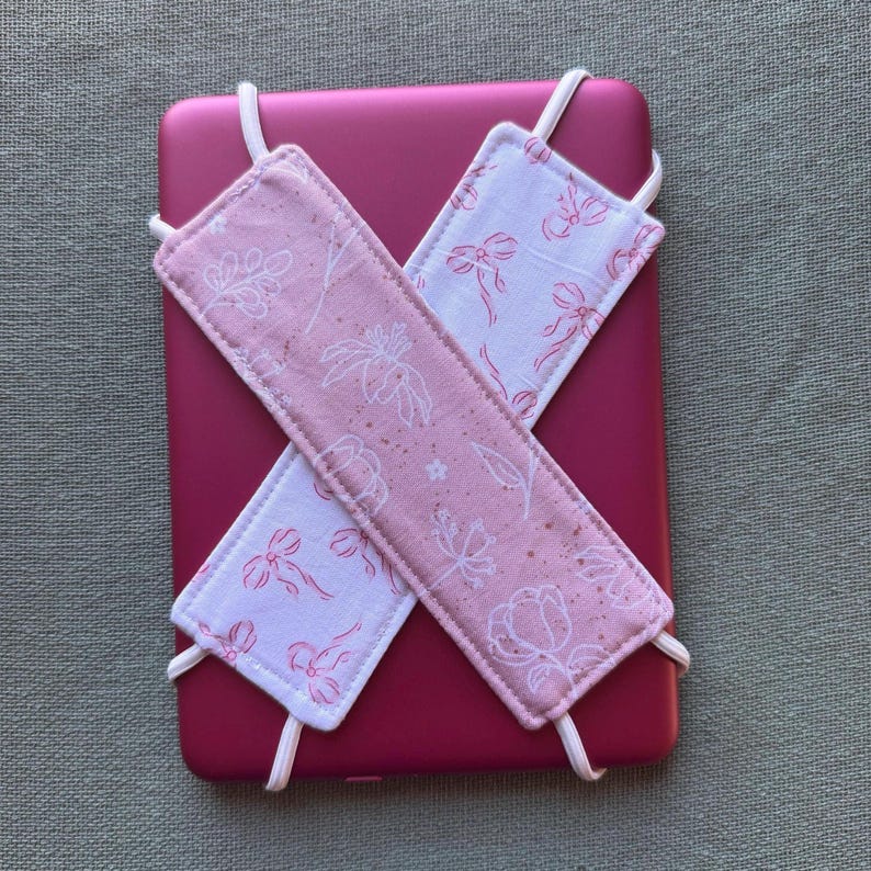 Op de afbeelding: Een magenta tablet met twee stoffen banden die kruisen. De roze en witte banden zijn versierd met bloemen- en strikpatronen. Witte elastische koorden bevestigen de banden aan de tablet en vormen een veilige houder.