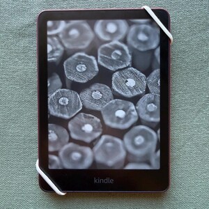 Op de afbeelding: Een Kindle e-reader met een donker scherm met een close-up van potloodpunten. Het apparaat heeft een donker frame met een roze-rode rand en een witte elastische band. Het woord "kindle" staat onderaan afgedrukt.