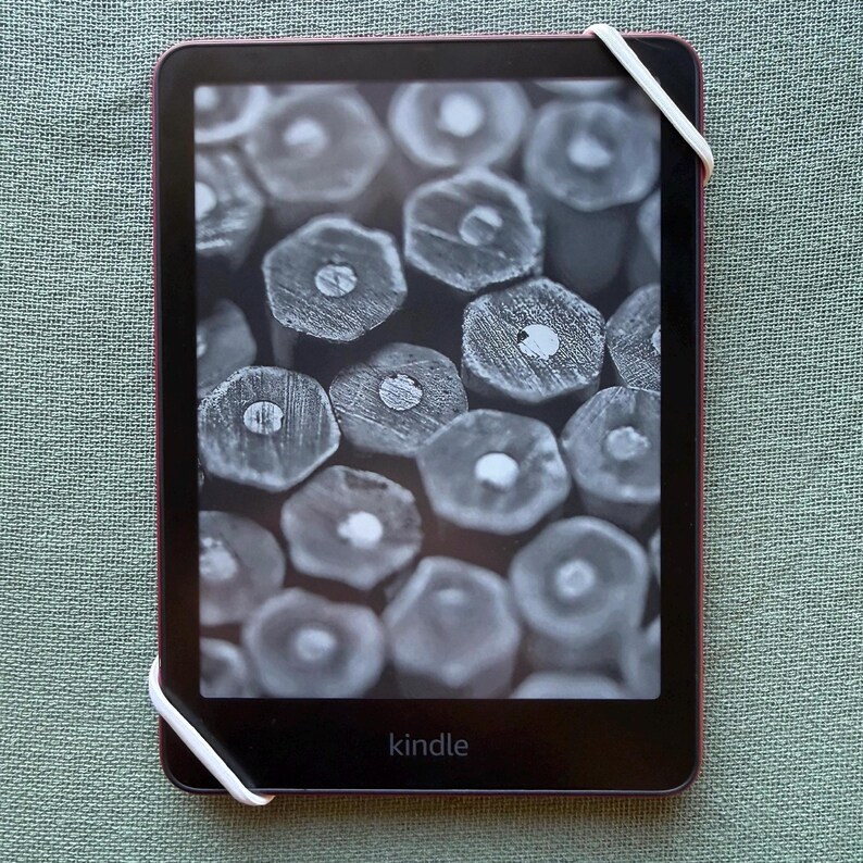 Op de afbeelding: Een Kindle e-reader met een donker scherm met een close-up van potloodpunten. Het apparaat heeft een donker frame met een roze-rode rand en een witte elastische band. Het woord "kindle" staat onderaan afgedrukt.
