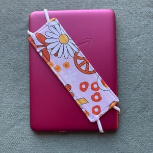 Op de afbeelding: Een roze e-reader met een bloemenbladwijzer. De bladwijzer heeft een madeliefje, een vredesteken en oranje en gele cirkels op een lichtroze achtergrond. Witte elastiekjes bevestigen de bladwijzer.