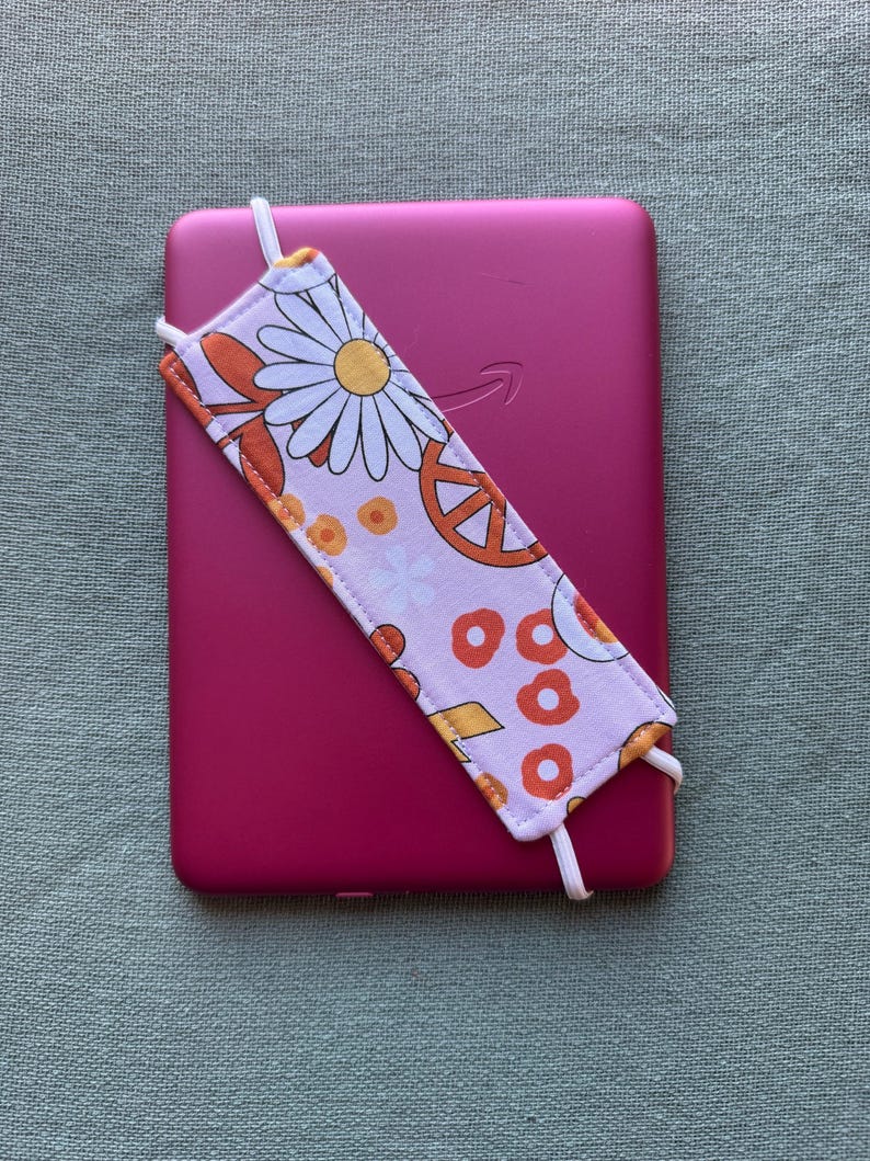 Op de afbeelding: Een roze e-reader met een bloemenbladwijzer. De bladwijzer heeft een madeliefje, een vredesteken en oranje en gele cirkels op een lichtroze achtergrond. Witte elastiekjes bevestigen de bladwijzer.