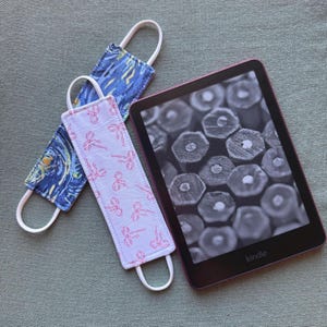 Op de afbeelding: Een e-reader met een zwart-wit afbeelding van potloden, naast twee stoffen bladwijzers. E&eacute;n bladwijzer heeft een blauw en geel sterrennachtontwerp, de andere een roze strikpatroon.