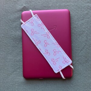 Op de afbeelding: Een magenta e-reader met een wit en roze strikpatroon bladwijzer. De stoffen bladwijzer heeft elastische banden om hem vast te zetten. Het Amazon-logo is zichtbaar op het apparaat. Afmetingen: ongeveer 17 cm.