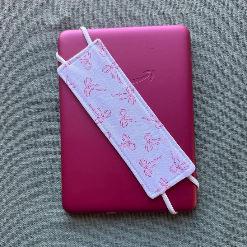 Op de afbeelding: Een magenta e-reader met een wit en roze strikpatroon bladwijzer. De stoffen bladwijzer heeft elastische banden om hem vast te zetten. Het Amazon-logo is zichtbaar op het apparaat. Afmetingen: ongeveer 17 cm.