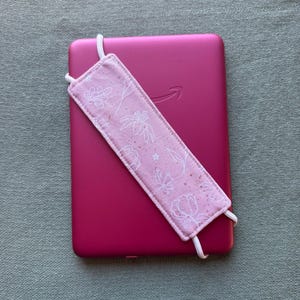 Op de afbeelding: Een roze e-reader met het Amazon-logo, gecombineerd met een bijpassende roze stoffen bladwijzer. De bladwijzer heeft een wit bloemenpatroon en is bevestigd met witte elastische koorden. De e-reader ligt op een getextureerd, lichtgroen oppervlak.