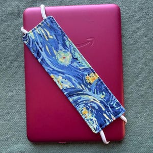 Op de afbeelding: Een magenta e-reader met een blauw en gele stoffen hoes met een sterrennachtontwerp. De hoes heeft witte elastische bandjes. Het Amazon-logo is zichtbaar op de e-reader.