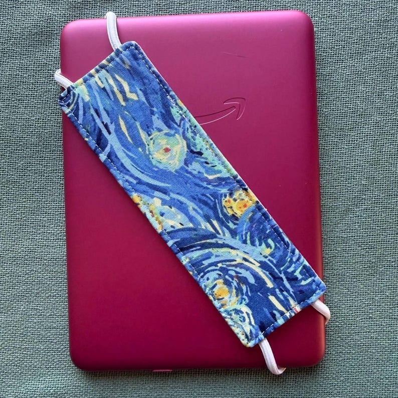 Op de afbeelding: Een magenta e-reader met een blauw en gele stoffen hoes met een sterrennachtontwerp. De hoes heeft witte elastische bandjes. Het Amazon-logo is zichtbaar op de e-reader.