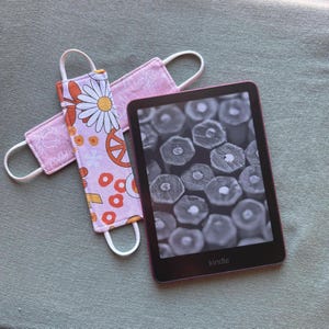 Op de afbeelding: Een Kindle e-reader met een zwart-wit scherm met een close-up van potloodpunten. Twee roze stoffen bladwijzers met witte elastische lussen liggen naast de e-reader. E&eacute;n bladwijzer heeft een madeliefje en vredesteken patroon.