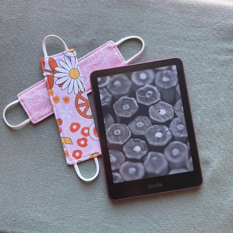 Op de afbeelding: Een Kindle e-reader met een zwart-wit scherm met een close-up van potloodpunten. Twee roze stoffen bladwijzers met witte elastische lussen liggen naast de e-reader. E&eacute;n bladwijzer heeft een madeliefje en vredesteken patroon.
