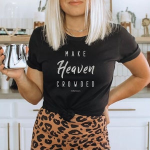 May include: Black t-shirt with the words "MAKE Heaven CROWDED" in white script. The shirt also has the hashtag "#LifeGoals" printed below the main text. The person is wearing leopard print leggings and holding a silver mug.
