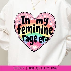 May include: A white sweatshirt with a rainbow heart design that says "In my feminine rage era".