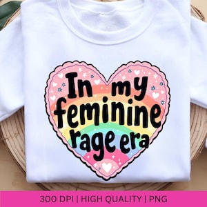 May include: A white sweatshirt with a pink heart design. The heart has a rainbow inside and the text "In my feminine rage era" in black.