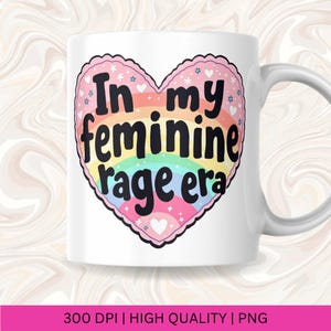 May include: White ceramic mug with a rainbow heart design. The heart has the text "In my feminine rage era" in black lettering.
