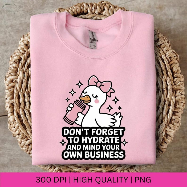 May include: A light pink sweatshirt featuring a cartoon duck with a pink bow, holding a pink drink. The text "DON'T FORGET TO HYDRATE AND MIND YOUR OWN BUSINESS" is below the duck. The sweatshirt is folded on a woven basket.